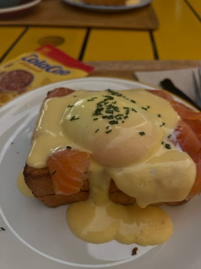A close-up, eye-level photograph presents a prepared dish of Eggs Benedict with smoked salmon on a white, round ceramic plate. Two poached eggs, one prominently centered, are thickly coated in a glossy, viscous yellow Hollandaise sauce. The sauce is garnished with finely chopped green chives. Beneath the eggs and sauce, multiple slices of orange-pink smoked salmon are visible, arranged over two pieces of golden-brown toast. Excess Hollandaise sauce has dripped onto the white plate. In the blurred background, a bright yellow tabletop is visible. A red and yellow packet labeled "Cola-Cao Original" is partially visible and out of focus in the upper-left portion of the frame, resting on the yellow table. A dark-colored fork is partially visible on the right side of the plate. Another out-of-focus dish is faintly discernible in the far upper-right background. The scene is set in an establishment in Santander, Spain, identifiable by the prominent Cola-Cao product, a Spanish brand.