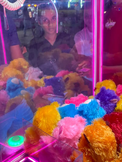The image shows a person reflected in the glass of a claw machine filled with colorful, fuzzy stuffed animals. The stuffed animals are various bright colors, including yellow, orange, pink, blue, and purple, and appear to be small, round, and bear-like. They are densely packed inside the machine, making it difficult to discern individual toys clearly. The machine has bright pink neon lights illuminating the top and sides. The reflection shows a person wearing a dark shirt, their face tilted and looking towards the viewer. The lighting in the machine is dim, with the neon lights being the most prominent light sources. The reflection of the person suggests a casual, indoor setting, possibly an arcade or amusement area. The overall impression is of a vibrant and playful environment, with the stuffed animals serving as the main objects of interest and the reflection adding a human element to the scene. The location cue of "Habaraduwa, Sri Lanka" does not provide any direct visual information in the image itself but suggests a tropical or subtropical setting where such entertainment venues might be found. There is text visible on a pink tag attached to one of the stuffed animals, which appears to be a logo or brand name, but it is too blurry to read.