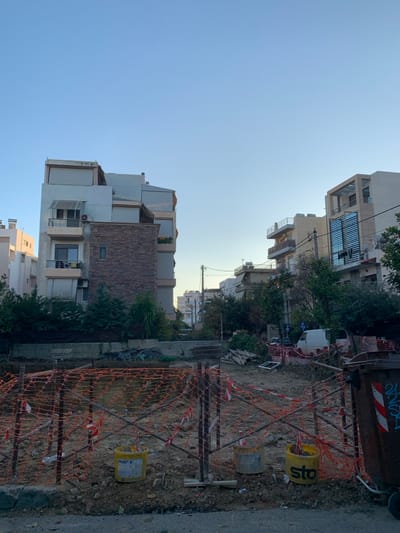 The image displays a sunny urban or suburban scene, likely in Marousi, Greece, under a clear blue sky. On the left side, several multi-story residential buildings line a street, featuring modern architecture with flat roofs, balconies, and mixed facades including light plaster and stone cladding. Lush green trees are visible between and in front of these structures.
The foreground and right side of the image reveal a dirt plot, enclosed by orange mesh construction netting supported by metal poles and concrete blocks. Within this fenced-off area, a discarded white window frame lies on the ground, and two yellow buckets are visible, one of which has the partial text "sto" printed on it. Further back on the right, a large dark dumpster is present, adorned with blue and red graffiti. A white van is parked on the street in the mid-ground, and utility poles with overhead lines extend across the scene.
The scene appears to be an active urban environment, possibly with an ongoing construction project or an undeveloped lot awaiting building. The time of day is bright daylight, indicated by the clear sky and strong illumination. No people are visible, and no specific event is captured beyond the static street and construction area.