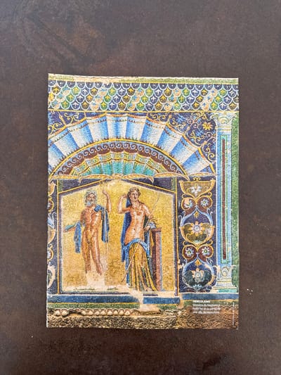 This media file is a postcard featuring a mosaic of Neptune and Amphitrite. The mosaic depicts two figures, presumably Neptune and Amphitrite, reclining on a golden background. The figures are rendered in warm, earthy tones of brown and tan, with blue drapery. They appear to be lying down, perhaps resting or in a scene of domestic tranquility.
The mosaic is framed by intricate decorative borders. On the left, there is a semi-circular fan-like pattern composed of blue, white, and yellow tesserae, reminiscent of a peacock feather or a stylized sun. This is bordered by a band of scalloped patterns in blue, green, and yellow. The top and right edges of the mosaic are also adorned with geometric patterns and bands of color, including blue, green, brown, and yellow. The right edge of the postcard shows a section of what appears to be a rough, textured wall, possibly the original setting of the mosaic.
Visible text on the postcard identifies the subject and location: "HERCULANO Mosaico de Neptuno y Anfitrite en la pared de una villa de Herculano." This indicates the mosaic is from Herculaneum, depicting Neptune and Amphitrite from a villa wall there. The location context provided, "l'Ametlla del Vallès, Spain," suggests this postcard may have originated from or been sent from this Spanish town. There is no indication of time of day or weather within the mosaic itself, and the emotional tone is serene.