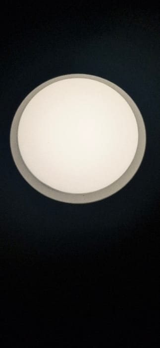 A close-up shot features a circular ceiling light against a dark background. The light has a distinct circular frame with a slightly rounded inner surface, suggesting a diffused glow. The color of the light is warm white or off-white, in stark contrast to the surrounding darkness. The image is simple, emphasizing the round light fixture and its gentle illumination.