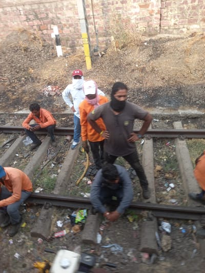 A group of people are working on a railroad track in Bhopal, India. There are at least seven individuals visible, some in orange shirts and others in varying colors. They are located on and around the railroad tracks. There is debris scattered around the tracks, and the area appears to be near a brick wall.