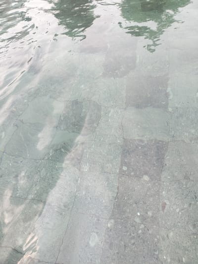The image displays a close-up, overhead view of a tiled surface submerged in clear water. The tiles appear to be stone or concrete, laid in a grid pattern, with visible grout lines. The water creates rippling reflections on the tile surface, distorting the view and creating abstract patterns of light and shadow. In the upper portion of the image, the reflection of foliage, likely from trees, is visible, appearing as green and darker shapes on the water's surface. The overall scene is serene and abstract, focusing on the interplay of light, water, and the textured tile beneath. The location is identified as Gunung Putri, Indonesia.