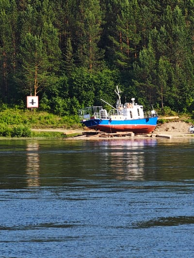 A blue and red vessel is beached on the shore of the Chulym River, adjacent to the city of Predivinsk, Russia. The vessel rests on a wooden support structure. A green forest borders the riverbank. A white and red sign with a black cross symbol is visible on the bank. The water reflects the surrounding trees.