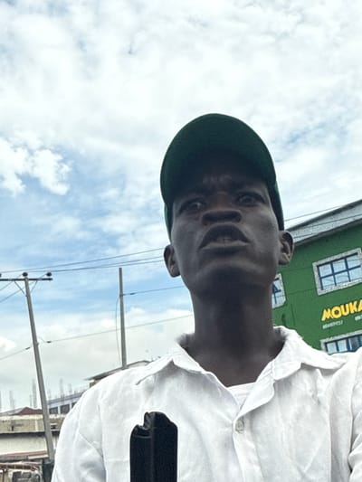 A daytime outdoor portrait shows a dark-skinned man, captured from a low angle, seemingly in Ojomu, Nigeria. He is wearing a dark green beret and a light-colored, likely white, collared shirt. The man looks directly at the camera with a neutral to subtle smiling expression. His face is partially shadowed, suggesting he is backlit.
The background features a bright, cloudy sky with scattered white clouds. To the upper right, a green building with multiple windows is visible. Yellow text on the building clearly displays "MOUKA" and below it, partially readable, is a phone number starting with "080". "MOUKA" is a prominent brand of mattresses in Nigeria. Utility poles with overhead wires are also visible in the lower portion of the background, indicating an urban or semi-urban environment. The image appears to be a casual photograph or selfie taken on a bright, possibly overcast, day.