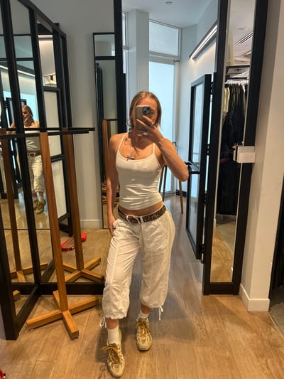A young woman takes a selfie in a retail environment, likely a clothing store, in Whistler, Canada. She is fair-skinned with blonde hair, wearing a white tank top, white capri pants with a studded brown belt, and beige sneakers. The woman is holding a smartphone in her left hand, presumably to take the picture, while her right hand rests on her waist. She is posing in front of a series of large mirrors, reflecting the clothing racks and interior design of the store. The reflections indicate a modern and bright space with wooden floors and clothing displays. Garments on hangers and other retail fixtures can be seen in the background.