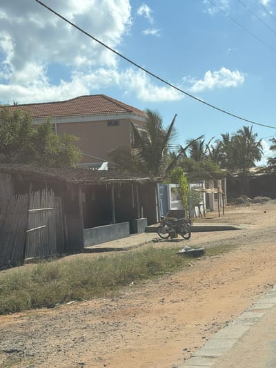 The image shows a street scene in Pemba, Mozambique. The road surface is dirt with a narrow strip of concrete along the edge. A bicycle is parked on the edge of the road in front of a building. The building is made of concrete and has a tiled roof. There is a fence made of bamboo or reeds next to the building. Palm trees are in the background, and the sky is blue with white clouds. Power lines stretch across the sky.