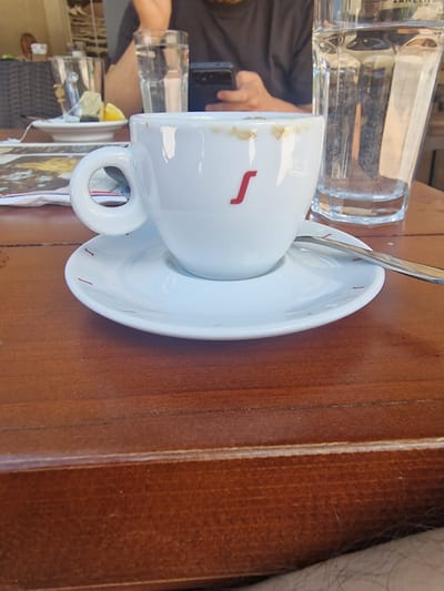 A close-up, eye-level shot focuses on a white coffee cup and saucer on a dark brown wooden table. The cup has a red stylized "S" logo. The saucer has small red dashes along its rim. A spoon rests on the saucer. Behind the cup, a glass of water, partially obscured by the cup, is visible. The background is blurred, showing a person in a black shirt holding a smartphone. In the further background, there are other tables, chairs, and a person in the distance, suggesting an outdoor seating area of a cafe. The scene is likely at a cafe in Cetinje, Montenegro, given the context.