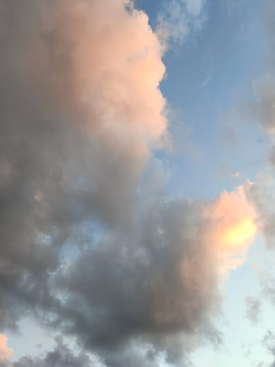 The image displays a cloudy sky. The clouds are a mix of dark grey and light pink and orange hues, suggesting either sunrise or sunset. The blue sky is visible in patches between the clouds. There are no discernible buildings, people, or objects in the scene. The image appears to be a photograph taken looking directly upwards at the sky.