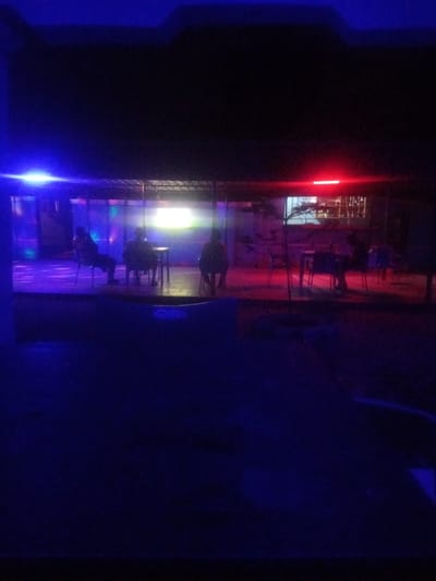 This image captures an outdoor seating area at night in Sabon Gari, Nassarawa, Nigeria. The scene is illuminated by blue and red neon lights, creating a moody ambiance. In the background, through an open-air structure, a bar or restaurant interior is visible with shelves stocked with bottles. Several individuals are seated at tables, appearing to be patrons. One person is seated on the left, facing away from the camera and towards the interior. Two other figures are seated at a table in the middle, also with their backs to the viewer. Further to the right, another person is seated at a table. The ground is paved or covered, and foliage is present in the middle ground. The overall impression is that of a lively nighttime establishment.