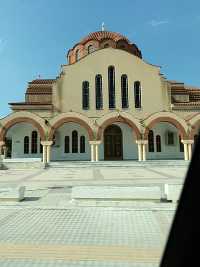 The image captures the exterior of a church in the Municipal Unit of Assos-Lechaio, Greece. The church features a dominant dome topped with a cross, constructed with red tiled roofing and brick accents. The facade is cream-colored with arched windows. Below the main structure, a series of brick arches supported by cream-colored columns create a covered walkway. The entrance is marked by a large wooden double door with intricate decorative patterns. The foreground shows a paved courtyard with geometric patterns in light-colored tiles and some concrete planters. The sky is clear and blue, suggesting a bright, sunny day. The view appears to be captured from a slightly elevated position, possibly from a vehicle, as indicated by the blurred foreground elements. There are no people visible in the image.