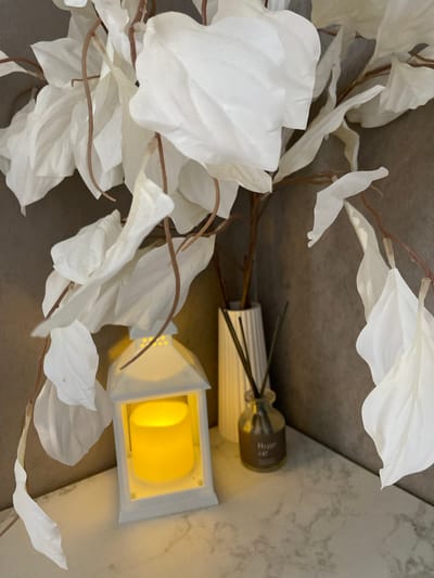 A high-angle shot shows a home interior scene with decor elements. Artificial white leaves on brown stems dominate the left side of the frame. To the right, an aroma diffuser with a small brown bottle and light-colored reeds sits next to a white lantern with a yellow, possibly artificial, candle inside. The background consists of surfaces with varying textures and colors, including a gray, potentially upholstered surface and a lighter, marbled texture. The location is not discernible from the image.