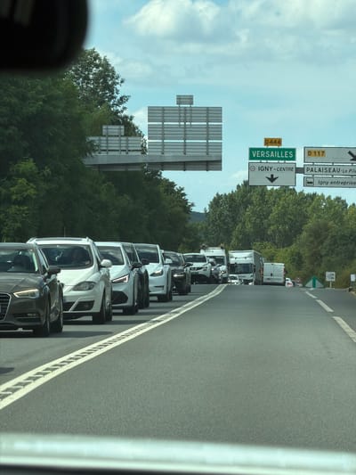 This media file shows a view of traffic on a highway in Palaiseau, France. The camera is positioned within a vehicle, looking forward at a long line of cars and trucks. The vehicles are traveling in a rightward direction, indicating a traffic jam. The road surface is asphalt, with white lane markings and a concrete barrier to the right. Trees and foliage line the side of the highway, suggesting a rural or suburban setting. Above the traffic, a bright blue sky with scattered clouds is visible.

Several road signs are present, offering directional information. One sign indicates "D 117" and "PALAISIEU-Le Pile". Another sign displays "D444 VERSAILLES" with an arrow pointing right, and "IGNY-CENTRE" also with a rightward arrow. The presence of these signs confirms the location as France.

The time of day appears to be daytime, with bright sunlight casting shadows. The weather seems to be clear and possibly warm, given the open sky and the presence of lush green trees. The overall mood conveyed is one of congestion and potential frustration due to the traffic standstill. The scene is dominated by the metallic surfaces of the vehicles and the natural elements of trees and sky.