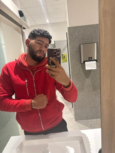 A male individual, approximately 20-35 years old, with dark curly hair and a beard, is depicted in a mirror selfie taken within a restroom or changing area located in the city of Radovljica, Slovenia. He is wearing a red zippered fleece hoodie over a red inner garment and dark pants. A black beaded bracelet is visible on his left wrist, with his left hand fisted near his hip. He holds a dark-colored smartphone, equipped with multiple rear cameras and a circular magnetic accessory, in his right hand, angled towards the mirror to capture the reflection. The foreground shows a white ceramic sink with a chrome drain. Background elements include a white overhead structure with a green and white "EXIT" sign in the upper left, a gray speckled tiled wall section to the right featuring a mounted silver automatic paper towel dispenser with a roll of white paper visible, and a light brown vertical panel or door frame on the far right. A faint reflection of a yellow notice or poster is visible on a white wall in the upper left background. The lighting is artificial.