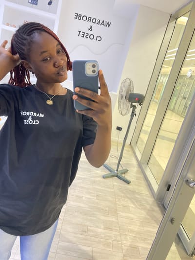 This media file depicts a young Black woman taking a mirror selfie in what appears to be a retail environment, possibly a clothing store, located in Unguwar Sarki, Nigeria. The woman is positioned centrally in the frame, holding a dark-colored smartphone to take the photo. She has braided hair with reddish-brown highlights and is wearing a black t-shirt with white text. A delicate necklace with a pendant is visible around her neck. The background shows a reflective surface, likely a large mirror, revealing more of the store.

Behind the woman and reflected in the mirror, a silver industrial fan on a stand is visible. The store's interior is characterized by light-colored tiled flooring and white shelving units, some of which contain clothing items. The overall lighting suggests it could be daytime, given the brightness reflecting from the ceiling lights and possibly natural light from an unseen window or entrance. The woman's expression is casual and relaxed. The text on her t-shirt reads "WARDROBE & CLOSET" in white, mirrored lettering. The overall impression is a candid snapshot of a shopping trip or a moment captured while browsing.