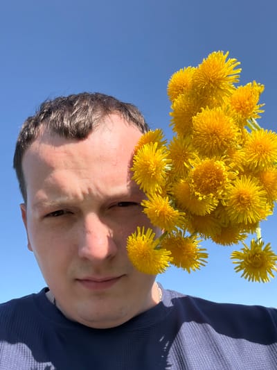 A low-angle shot of a man, possibly in his late 20s or early 30s, holding a bouquet of bright yellow dandelions against a clear blue sky. The man's face is prominently featured, with short dark hair, light skin, and dark eyes. He is wearing a dark blue long-sleeved shirt. The image appears to be taken outdoors, possibly in the city of Кемуль, Russia, as indicated by the instructions. The location is implied by the absence of specific landmarks or distinguishing features. The focus is on the man and the dandelions, with the sky serving as a backdrop.