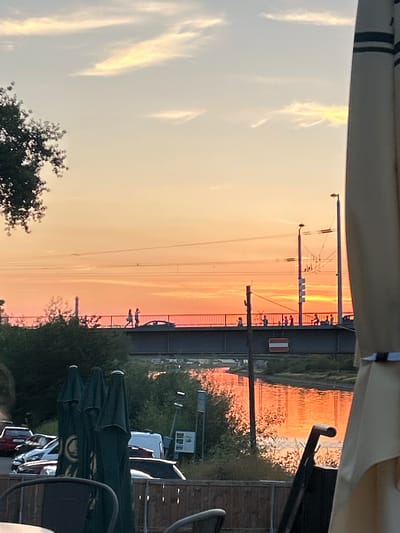 The image captures a serene outdoor scene at sunset, likely in Mannheim, Germany. The sky is painted with warm, vibrant hues of orange and yellow, indicating the sun is low on the horizon. Wispy clouds catch the fading light, adding a painterly effect to the sky.
Dominating the mid-ground is a bridge, possibly a railway bridge, with visible overhead electrical lines and supports. A few silhouetted figures can be seen walking on or near the bridge, suggesting a public walkway. The water of a canal or river reflects the warm colors of the sunset, creating a shimmering surface.
In the foreground, partially obscuring the view, is the edge of an awning or canopy. Below this, several dark green, closed umbrellas are stacked, hinting at an outdoor seating area, perhaps a café or restaurant that is preparing to close for the day or is not yet open. A glimpse of parked vehicles, including a white van and other cars, can be seen on the right side, suggesting proximity to a road or parking area. Trees and green foliage add to the natural elements of the scene.
The overall atmosphere is peaceful and tranquil, typical of a calm evening by the water as day transitions into night. The setting appears to be an urban or suburban area, given the presence of infrastructure like the bridge and power lines, combined with natural elements and the outdoor seating area. No legible text is visible in the image.