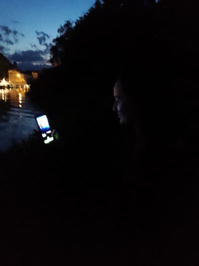 The image depicts a person's profile in silhouette, facing right, with their face illuminated by the glow of a flip phone they are holding. The phone is open and emitting a bright blue and green light. The person appears to be sitting or standing near a body of water, possibly a lake or river, with the water reflecting lights from buildings in the background. The sky above is a deep twilight blue with some clouds. The overall scene is dark, with most of the environment obscured by shadow. The hint images suggest the location may be in Schwerin, Germany, potentially near a waterfront area with buildings visible in the distance.