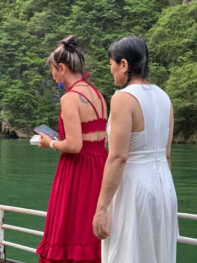 Two women stand next to each other on what appears to be a boat or platform with a white metal railing, against a backdrop of lush green vegetation and water. The woman on top is wearing a white dress. Her hair is black and pulled back. The woman on the bottom is wearing a red dress with a ruched back detail. She holds a smartphone and a small white object in her hands. She is also wearing a white smartwatch and blue tassel earrings, and her hair is pulled up in a bun. The picture was taken in Đồng Văn Commune, Vietnam.