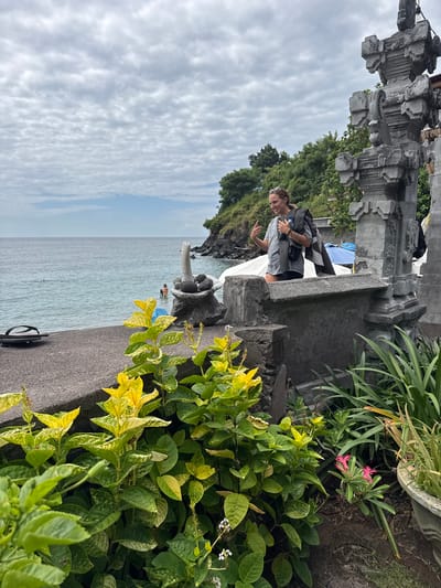 In Abang, Indonesia, a woman stands near a concrete wall, holding a gray and black striped towel over her shoulder and a black bottle in her right hand. She is wearing a gray t-shirt and black shorts. She is gesturing with her left hand, with her index and pinky fingers extended, and looking down. In the background, the ocean is visible with several people in the water. A rock formation with a carved stone sculpture is to her right. In the foreground, lush green and yellow foliage covers a stone wall. The sky is overcast with a textured pattern of clouds.
