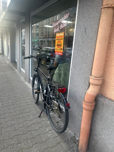 A black bicycle is parked on a cobblestone sidewalk in front of a store with a large glass window. The store displays a sign that reads "Hausgemachter Steak Döner". A bright yellow flyer is attached to the window, advertising "UNIKAT DÖNER !BRANDNEU! !DÖNERBURGER!" with a price of "3,90". The bicycle has a basket and a red taillight. A brown downspout runs vertically along the grey textured wall of the building. In the reflection of the window, several cars and buildings are visible, suggesting an urban environment. The scene appears to be in Gelsenkirchen, Germany, based on the style of the buildings and signage. There are no people clearly visible in the image. The lighting suggests it is daytime, possibly late afternoon due to the angle of the sun.