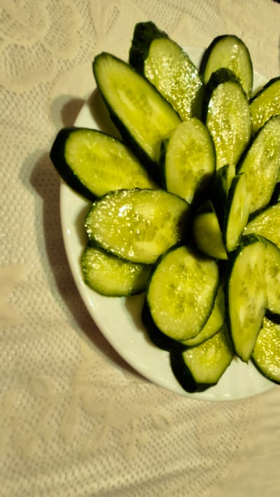 A white plate is filled with thinly sliced cucumbers on a textured tablecloth. The cucumbers are a vibrant green with visible seeds and a slight sheen, suggesting they may have been recently dressed or prepared. The slices are arranged in a somewhat overlapping pattern, creating a layered effect.

The setting appears to be indoors, likely a dining area, given the presence of the plate and tablecloth. The lighting suggests it could be evening or a dimly lit room, as there are subtle highlights and shadows on the cucumbers. The tablecloth has a lace-like pattern, adding a decorative element to the scene. No people are visible in the image. The focus is entirely on the prepared cucumbers. There is no visible text in the image. The context of Ialoveni, Moldova, does not provide specific visual cues within the image itself, but suggests a potential domestic or culinary context in that region.