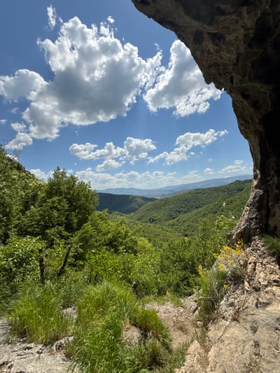 The photograph depicts a view from inside a cave in the Lipnitsa area of Bulgaria. The cave entrance frames a landscape of forested hills and valleys, with a blue sky dotted with white clouds visible through the opening. The cave walls are rough and rocky, with some vegetation growing near the entrance. The overall scene suggests a natural, mountainous environment.