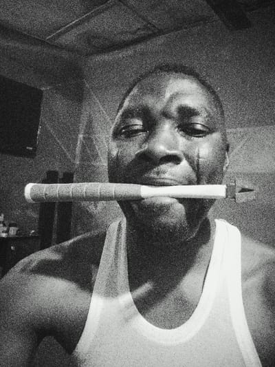 A grainy, black and white image displays an adult male in an indoor setting within Gawuna, Nigeria. The subject, positioned centrally and facing forward, wears a light-colored tank top. Prominent vertical scars are visible on both cheeks. He holds a slender, approximately 12-inch long object horizontally in his mouth, clamped between his teeth. The object features a white handle section with dark, transverse bands and tapers to a small, triangular fletching-like end on the right. To the upper left of the subject, a wall-mounted television is partially visible, with indistinct objects resting on a surface below it. The ceiling is visible at the top of the frame. The image exhibits significant visual noise.