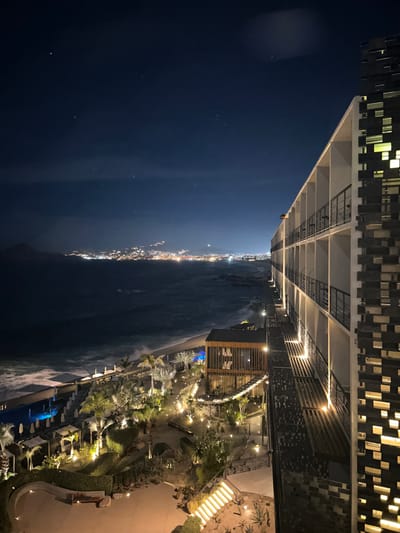 The image depicts a night scene in Cabo San Lucas, Mexico. In the foreground, a multi-story building with illuminated windows is seen from an elevated perspective. The building's modern architecture is visible, with a patterned facade and structural elements. Below, a landscaped area featuring illuminated pathways, palm trees, and other vegetation is present. A body of water, presumably the ocean, separates the illuminated resort area from a coastline dotted with city lights. The night sky is dark, with a few visible stars. The overall scene suggests a high-end resort or hotel overlooking the coastline of Cabo San Lucas.