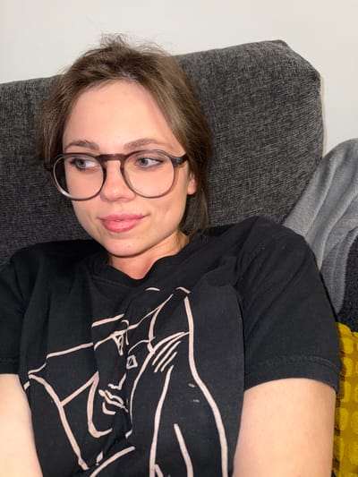 A young woman with light brown hair and glasses is lying on a dark gray sofa. She is wearing a black t-shirt with a white graphic design. Behind the sofa are a light gray blanket and a yellow pillow. The background is a white wall. The image's location in Balatonlelle, Hungary, is not discernible.