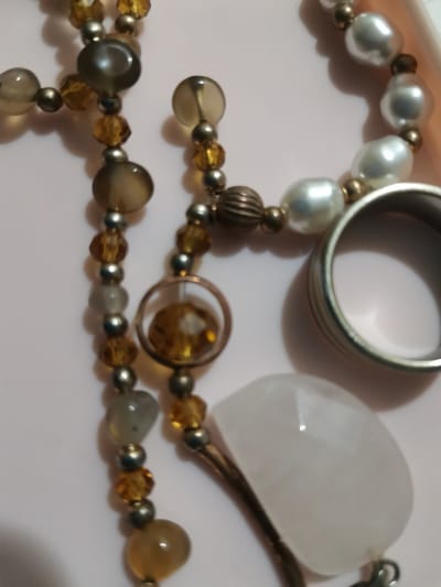 The image shows a close-up of several pieces of jewelry arranged on a light pink surface, possibly a table. The jewelry includes a necklace with various beads, some appearing to be pearls, amber-colored crystals, and opaque grey or brown stones. There is also a silver-toned ring with textured detailing and a pendant with a clear, faceted stone, possibly quartz or glass, and a copper-colored frame. The overall impression is of a collection of decorative accessories. There are no people, actions, or interactions depicted, and the environment is a plain, unadorned surface. The image does not provide any information about the location in Songjiang District, China.