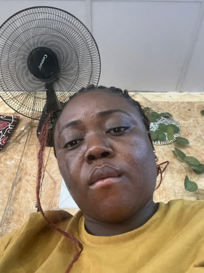 An individual is depicted in an indoor selfie, centered in the frame, looking directly at the camera. The subject is an adult female with dark skin, dark braided hair, and a small silver nose piercing on her right nostril. She is wearing a mustard yellow long-sleeved top. A pair of reddish-brown framed glasses are visible behind her right ear. In the upper left background, a black Century brand standing fan is partially visible. The wall behind the subject is covered with light brown or beige textured tiles. A decorative item with red, black, and white swirl patterns is partially visible on the left side of the wall, and a green, leaf-like wall decoration is visible on the right side. The ceiling is white or light grey. The setting is an unidentified indoor space within the city of Abuja, Federal Capital Territory, Nigeria, consistent with a residential or commercial interior. No specific street address or landmark is discernible.