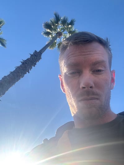A man with light skin and graying hair takes a low-angle selfie against a bright blue sky. He is positioned above a tall palm tree with a shaggy trunk and fan-shaped leaves. Sunlight flares intensely in the lower right corner of the image, obscuring some details. The man's face shows lines and a somewhat neutral expression.