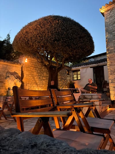 In the city of Kassandra Municipal Unit, Greece, at dusk, an outdoor seating area is depicted. Two men are visible, seated on chairs. One man, wearing a dark shirt, is seated on a bench in front of a stone wall. The other man, wearing a green shirt and glasses, is seated in a wooden chair on a raised stone platform, holding a mobile phone. In the foreground, empty wooden chairs are arranged. A large, rounded tree with dark foliage is centrally positioned. The surrounding architecture features stone walls and tiled roofs, illuminated by warm artificial lighting. The scene suggests a relaxed evening ambiance.