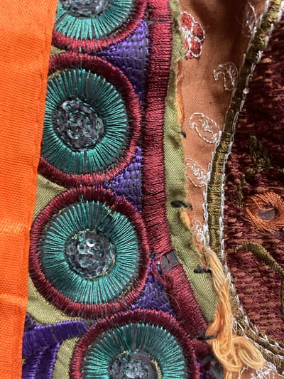 The image is a close-up, overhead shot of an orange fabric textile with intricate embroidery in various colors, including teal, purple, red, green, and brown. The embroidery features concentric circular patterns, some with sequins, and decorative stitching. There are also some frayed threads and seams visible, suggesting it might be a piece of clothing, a tapestry, or a decorative element. The image appears to be taken indoors, with natural light. There is no identifiable location in Sabadell, Spain, or any specific details about people or actions.