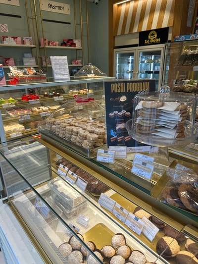 The image depicts a pastry display case inside a bakery, possibly "Le Goût" in Kolašin, Montenegro, based on the sign visible in the background. The case contains various pastries and cookies, including those labeled as part of a "Posni Program" (Lenten program). The pastries are displayed under clear domes and in plastic containers, with price tags visible for items such as "Kuluri dzem" (€0.30), "Žerbo," "Bajadera" (€1.70), "Išleri" (€0.80), "Američki Cookies" (€0.70), and "Padobranac" (€0.70). Other baked goods are visible, like chocolate-covered cookies and coconut-covered treats. Shelves with products are visible in the background of the bakery. A sign reading "PASTRY" hangs to the left.
