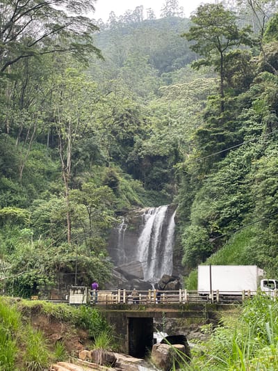 The image captures the Ramboda Falls in Sri Lanka. A waterfall cascades down a rocky cliff, surrounded by dense green vegetation. A man wearing a purple shirt stands on a metal bridge-like structure that appears to be a part of a water management system, potentially a small dam. Adjacent to the structure is a white truck. The scene is set in a lush, mountainous environment, typical of the Ramboda region.