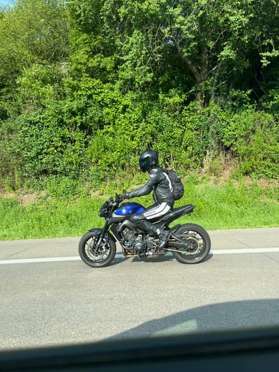 A motorcyclist in full black and white leather gear is leaning into a curve on a black and blue motorcycle on an asphalt road with a white dividing line, with lush green vegetation bordering the roadside. The photo appears to have been taken from a vehicle. The specific location in Bedburg, Germany cannot be determined from the image alone.