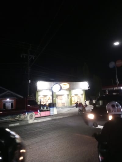 The image depicts a night scene in Walennae, Indonesia, specifically on Jalan Poros Walennae. A red flatbed truck is parked on the side of the road, facing left. In front of the truck, a building with a bright, illuminated sign is visible, appearing to be a commercial establishment. Several motorcycles are on the road, with riders visible. The road surface is asphalt, and there are power lines overhead supported by a utility pole. The overall atmosphere is that of a bustling street at night.