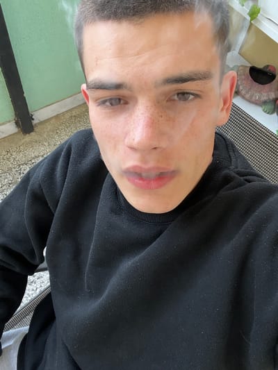 A close-up, rotated perspective selfie depicts a young Caucasian male with short, graying hair. He has light eyes, freckles, and a fair complexion. He wears a black crew-neck sweatshirt. The background reveals a green painted surface, and a patterned chair. The location and city are not Bulgaria or Sopot.