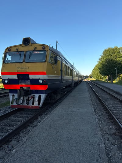 The image shows a yellow and blue train with the number DR1AM-222.4 printed on its front, parked on a railway track. The train has a distinctive red, white, and blue stripe pattern on its front. The setting appears to be a train station in Augšlīgatne, Latvia. The station platform is visible next to the train, made of grey gravel. On the right side of the image, another set of parallel railway tracks runs into the distance, bordered by a line of lush green trees. The sky is clear and blue, indicating a bright day. The overall scene is static and peaceful, with no people or activity visible.