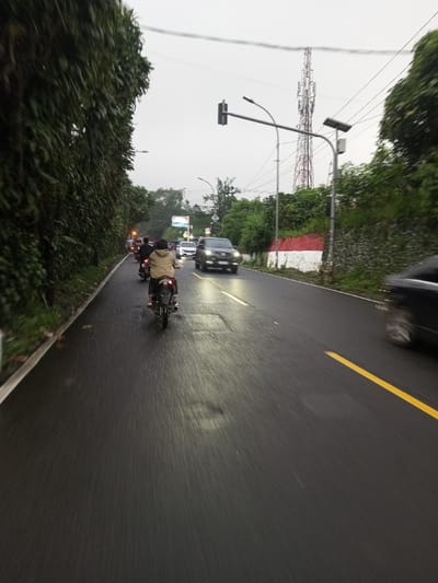 This image captures a street scene in the city of Dawathang_Dorjibi_ Kashingtsawa, Bhutan, under overcast daylight conditions. The paved asphalt road appears wet, reflecting ambient light.

The road features a solid white line on the far left edge and a solid yellow line marking the right boundary of the visible lane. Two distinct rectangular patches of darker asphalt indicate repairs on the road surface.

On the left side, a dense, tall wall of dark green foliage extends upwards, bordering the road. On the right side, a retaining wall painted with horizontal red and white stripes supports a vegetated embankment. Above the embankment, a tall, metallic lattice telecommunications tower is prominent against the gray sky.

Overhead infrastructure includes a street light fixture mounted on a pole on the right, equipped with a solar panel. Further down the road, a traffic light assembly hangs from an overhead gantry. Multiple utility lines traverse the upper portion of the frame.

Traffic is present in both directions. In the foreground, a person wearing a light-colored jacket is riding a dark motorcycle away from the camera in the left lane, with the red taillight illuminated. At least one additional motorcycle is visible behind the primary motorcycle. In the opposing lane, a dark-colored SUV with illuminated headlights is approaching. Other vehicles, including cars and motorcycles, are visible further in the distance. A dark vehicle appears blurred in motion on the far right edge of the foreground, moving from right to left.