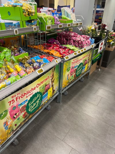 The image shows two metal supermarket displays, both filled with Easter-themed products and decorated with signs featuring the German phrase "Bis zu 75 Fröhliche Osterzeit Artikel" which translates to "Up to 75 Happy Easter Time Articles". The displays are located in a store in the city of Köln, Germany, at coordinates 50.919 latitude and 6.923 longitude. The displays are arranged in a row, with a grey tiled floor between them. The leftmost display features a variety of chocolate Easter eggs, wrapped in foil and plastic, arranged in rows. There is a smaller display on the left, featuring yellow and green cardboard boxes with images of chocolate bunnies on them. The rightmost display features a similar assortment of chocolate Easter eggs, as well as chocolate bunnies and other Easter-themed candies, all individually wrapped. The displays are lit by fluorescent lights overhead, and the background is slightly blurred.