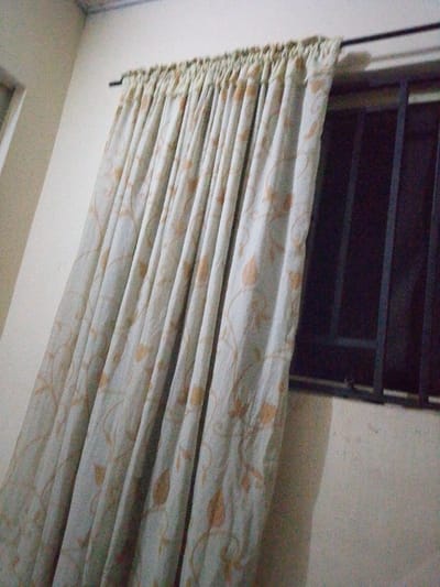 The image captures an interior scene in Abatete, Nigeria, focusing on a window and curtain assembly. A light-colored curtain, featuring a repeating pattern of light brown or tan foliage, hangs from a thin, black metal curtain rod mounted on a light-colored wall. The curtain is pleated and extends diagonally across the frame from the upper left to the lower right. To the immediate right of the curtain, a portion of a window is visible, fitted with dark-colored metal security bars arranged in a grid pattern. Additional light-colored wall sections are present to the left of the curtain and below the window. A small segment of a white ceiling with a decorative trim is visible in the upper left corner. No individuals or dynamic actions are depicted.