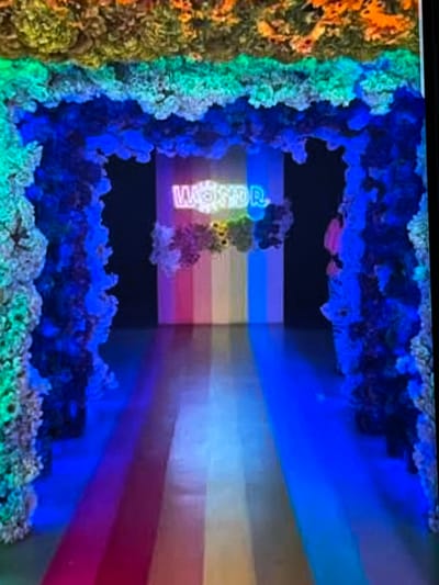 The image depicts an indoor art installation with a vibrant and surreal aesthetic. The primary subject appears to be a tunnel-like structure or an alcove, framed by an organic, coral-like material that glows with various colors, predominantly blues and greens, with hints of orange and yellow on the left side. This material has a textured, almost rocky appearance.
Inside the framed space, the background is a dark void. Emerging from this darkness and extending into the foreground are horizontal stripes of color, creating a sense of depth and a rainbow effect. These stripes range from yellow and light blue to purple and pink.
Illuminated within the dark void, centered within the colorful stripes, is an object that appears to be a neon sign or a light sculpture. The visible text on this object reads "WORLDP". The letters are white and outlined in a lighter color, and they are partially obscured by the colorful, organic framing material. The overall lighting is dramatic and artificial, with the colors creating a dreamlike or otherworldly atmosphere.
The scene does not feature any people. It is difficult to ascertain the time of day or weather conditions as it is an indoor installation. The location context of Nijmegen, Netherlands, does not provide specific visual cues within the image itself. The notable details are the striking use of color, the contrasting textures of the organic framing and the smooth stripes, and the partially visible text. The overall emotion evoked by the image is one of wonder and intrigue due to the unusual and immersive visual presentation.