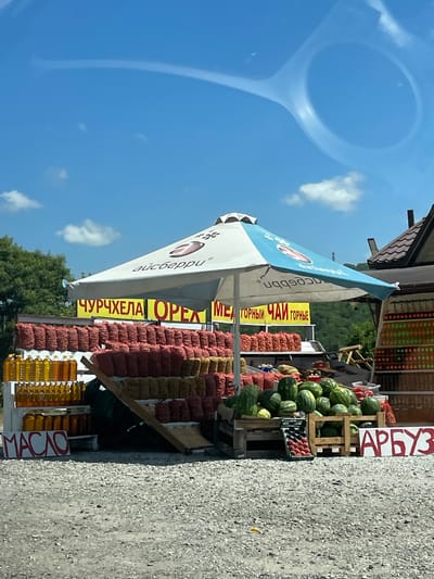The image depicts an outdoor market stall in Dzhubga, Russia, on a sunny day with a clear blue sky and scattered clouds. The stall is covered by a large white and blue umbrella. In front of the stall, rows of bags filled with what appear to be nuts or dried fruits are displayed on tiered shelves. To the left, several bottles of yellow liquid, likely cooking oil, are arranged in stacks. Further to the right, a display of watermelons and other produce is visible, along with a crate of red fruits, possibly peaches. Two signs are prominently displayed: one reads "MASLO" (oil) in red letters on a white background, and another sign on the right side reads "АРБУЗ" (watermelon) in large red letters on a white background. Behind the stall, a building with a tiled roof is partially visible, and a lush green hillside with trees forms the backdrop. The ground in the foreground is covered with gravel. There are no people visible in the image.