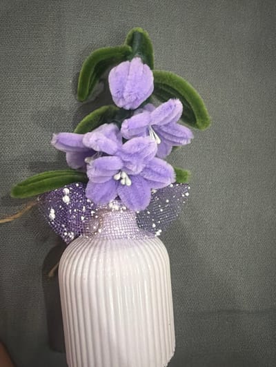 A close-up, eye-level shot captures a decorative arrangement against a dark grey, textured background. The primary subject is a vertical white, ribbed bottle adorned with artificial flowers and ribbons.
The bottle, appearing to be made of ceramic or opaque glass, features distinct vertical fluting along its visible length. Attached to the upper neck of the bottle are several artificial flowers and leaves. The flowers are soft, plush, and light purple (lavender) in color, with small white bead-like stamens clustered in their centers. At least five individual flower blossoms are visible, arranged in a cluster. Interspersed among the flowers are several artificial green leaves, also appearing to be made of a soft, velvety material.
Beneath the flower arrangement, a decorative element is tied around the bottle's neck. This consists of a wide, sheer, purple mesh ribbon, speckled with small white dots, fashioned into a bow-like shape. A thinner, solid purple ribbon is partially visible underneath the mesh. A thin, light brown string or twine extends from the left side of the arrangement, suggesting a hanging mechanism.
The background is uniform and featureless, composed of a dark grey material with a fine, consistent texture, possibly fabric or carpeting. There are no identifiable landmarks, people, or other objects to determine a specific geographical location. Based on the visual evidence, it is not possible to determine a location in the city of Vladaya, Bulgaria, or any other specific city or country.