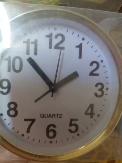 This is a close-up, slightly angled shot of a round analog clock with a gold-colored rim, possibly a wall clock or a clock that was part of another item. The clock face is white with black numerals and markers. The hour hand is pointing to approximately the 10 o'clock position, and the minute hand is pointing towards the 11 o'clock position, indicating the time is roughly 10:55. There is also a thin second hand visible, pointing towards the 5 o'clock position. The word "QUARTZ" is printed vertically in black text on the right side of the clock face. The clock is enclosed in clear plastic packaging, with some reflections and glare on the surface, suggesting it is new or being stored. The background is out of focus, appearing to be a cluttered indoor setting with warm, earthy tones. There are no people or animals visible. The scene is static, with no activity taking place beyond the movement of the clock's hands. The lighting is bright and somewhat diffused. The location context of Marynivka, Ukraine is noted, but there are no specific visual cues in the image to confirm this location.