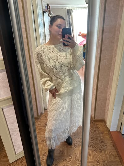 In the mirror reflection, a young woman stands in a room, holding a smartphone to take a picture of herself. She is wearing a cream-colored, crocheted sweater with a wide neckline and puffy sleeves, paired with a long, white, textured skirt. Her dark hair is pulled back into a ponytail, and she looks directly at the phone with a neutral expression.
The background of the reflection reveals a room with patterned wallpaper and linoleum flooring. Sunlight streams in from a window, casting bright light and creating shadows. A glimpse of clothes hanging in the distance suggests the room might be a bedroom or a dressing area. The overall setting appears to be a domestic interior, likely in Yaroslavka, Russia. The image is taken at an angle due to the reflection in the mirror. No visible text is present.