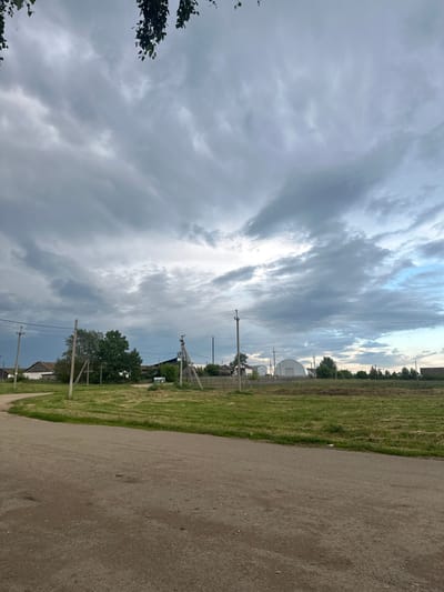 The image depicts a rural scene in Yaroslavka, Russia, on a cloudy day. A dirt road curves from the bottom left of the frame into the middle ground. To the left of the road, a grassy field stretches towards a small cluster of houses. Behind the houses, a line of trees is visible. In the middle ground, several utility poles with power lines traverse the scene. Further in the distance, a large, arched structure, possibly a greenhouse or agricultural building, stands out amongst more trees and houses. The sky is dominated by dramatic, grey, and white clouds, with patches of lighter blue sky visible towards the right. The overall impression is one of a quiet, overcast day in a village setting.